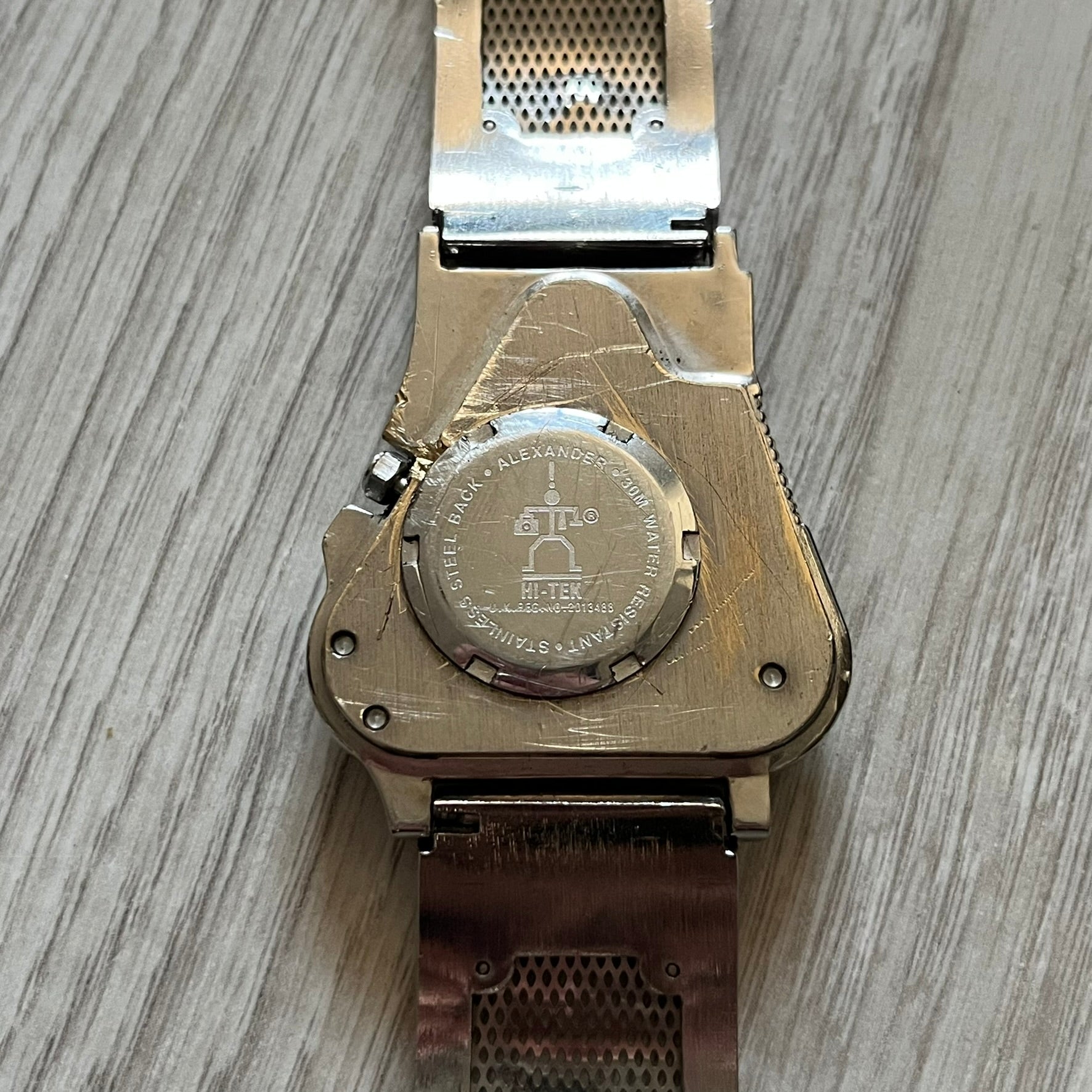 The Hi-Tek Alexander “The Engine” analog watch is a 1990s model made in England, designed with a sculptural case shaped like a motorcycle engine. It features a black dial with Alexander Hi-Tek figure logos in place of hour markers, pink hour and minute hands, and a Hi-Tek figure logo used as the seconds hand. A small sub-section at the bottom displays the day in numeric form, while visible screws and textured detailing are integrated into the engine-style face.&nbsp;The watch has an alloy case with a stainl
