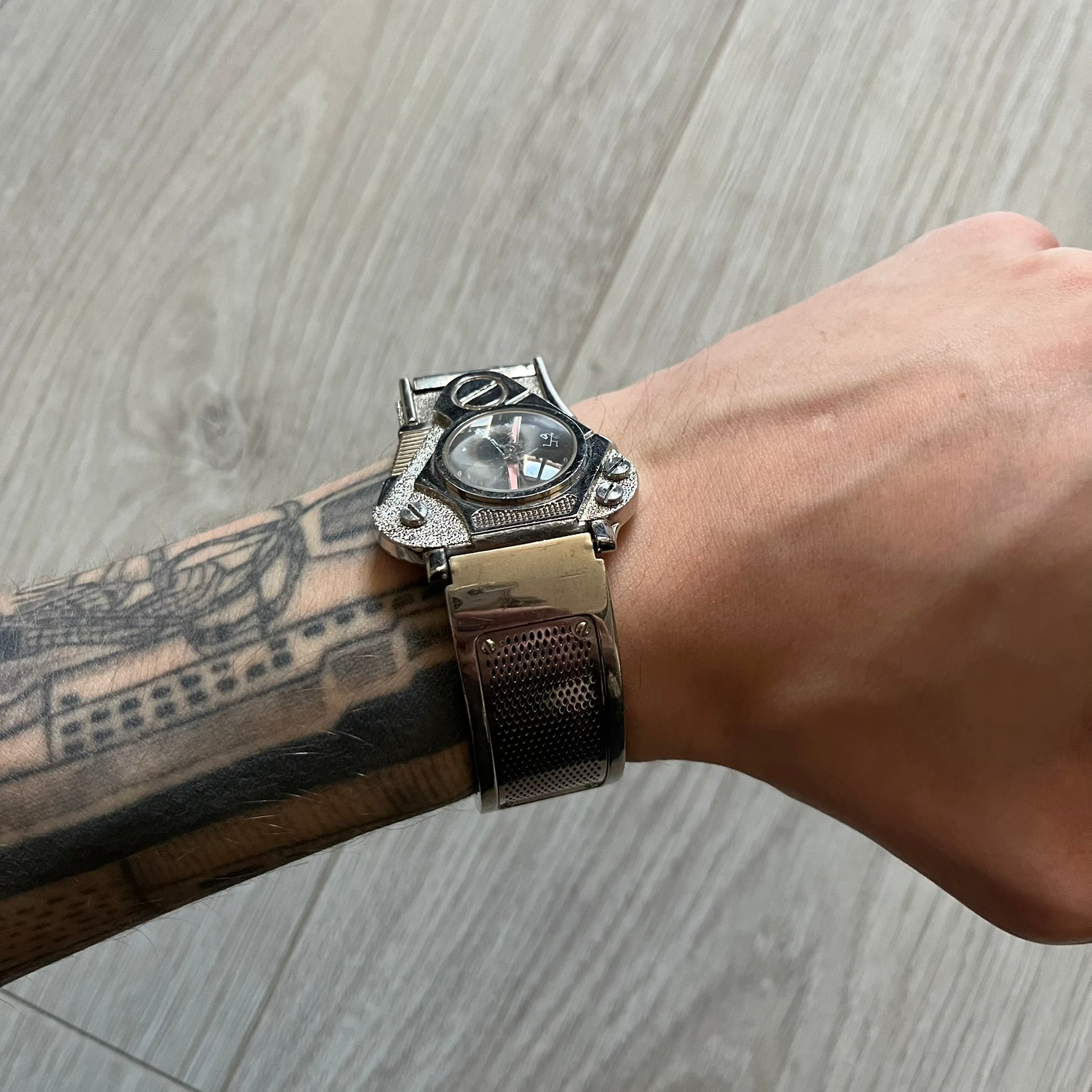 The Hi-Tek Alexander “The Engine” analog watch is a 1990s model made in England, designed with a sculptural case shaped like a motorcycle engine. It features a black dial with Alexander Hi-Tek figure logos in place of hour markers, pink hour and minute hands, and a Hi-Tek figure logo used as the seconds hand. A small sub-section at the bottom displays the day in numeric form, while visible screws and textured detailing are integrated into the engine-style face.&nbsp;The watch has an alloy case with a stainl