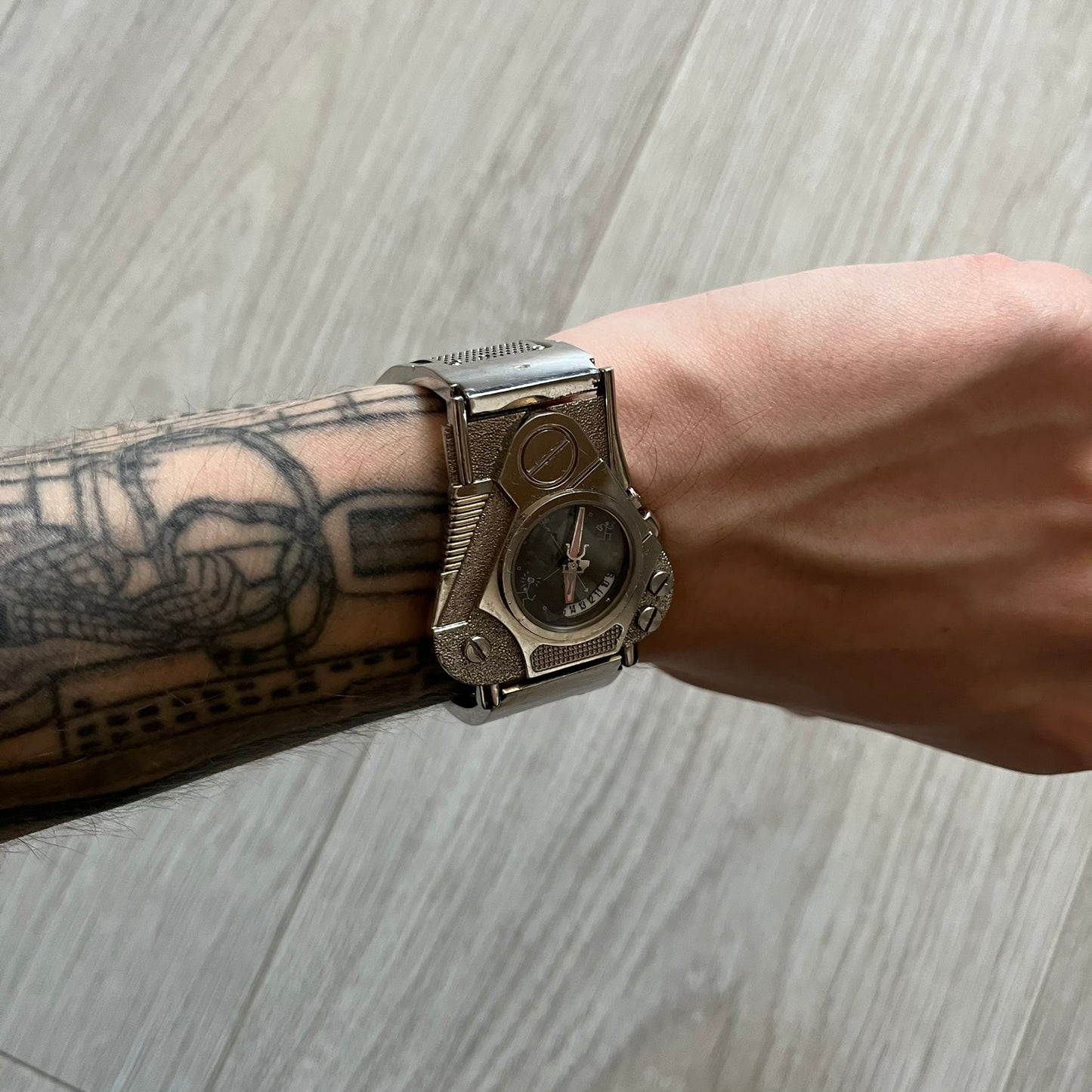 The Hi-Tek Alexander “The Engine” analog watch is a 1990s model made in England, designed with a sculptural case shaped like a motorcycle engine. It features a black dial with Alexander Hi-Tek figure logos in place of hour markers, pink hour and minute hands, and a Hi-Tek figure logo used as the seconds hand. A small sub-section at the bottom displays the day in numeric form, while visible screws and textured detailing are integrated into the engine-style face.&nbsp;The watch has an alloy case with a stainl
