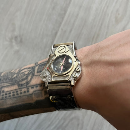 The Hi-Tek Alexander “The Engine” analog watch is a 1990s model made in England, designed with a sculptural case shaped like a motorcycle engine. It features a black dial with Alexander Hi-Tek figure logos in place of hour markers, pink hour and minute hands, and a Hi-Tek figure logo used as the seconds hand. A small sub-section at the bottom displays the day in numeric form, while visible screws and textured detailing are integrated into the engine-style face.&nbsp;The watch has an alloy case with a stainl