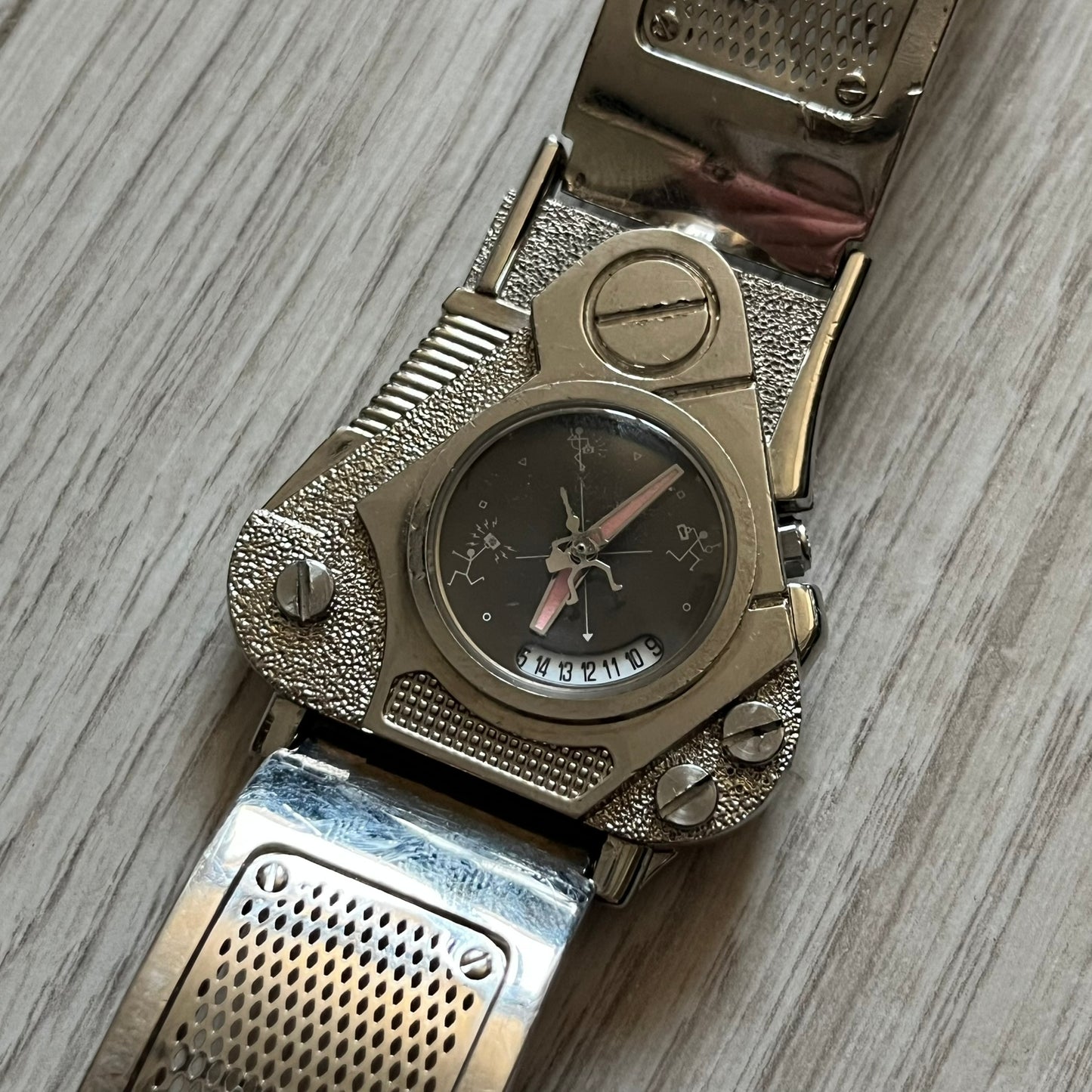 The Hi-Tek Alexander “The Engine” analog watch is a 1990s model made in England, designed with a sculptural case shaped like a motorcycle engine. It features a black dial with Alexander Hi-Tek figure logos in place of hour markers, pink hour and minute hands, and a Hi-Tek figure logo used as the seconds hand. A small sub-section at the bottom displays the day in numeric form, while visible screws and textured detailing are integrated into the engine-style face.&nbsp;The watch has an alloy case with a stainl