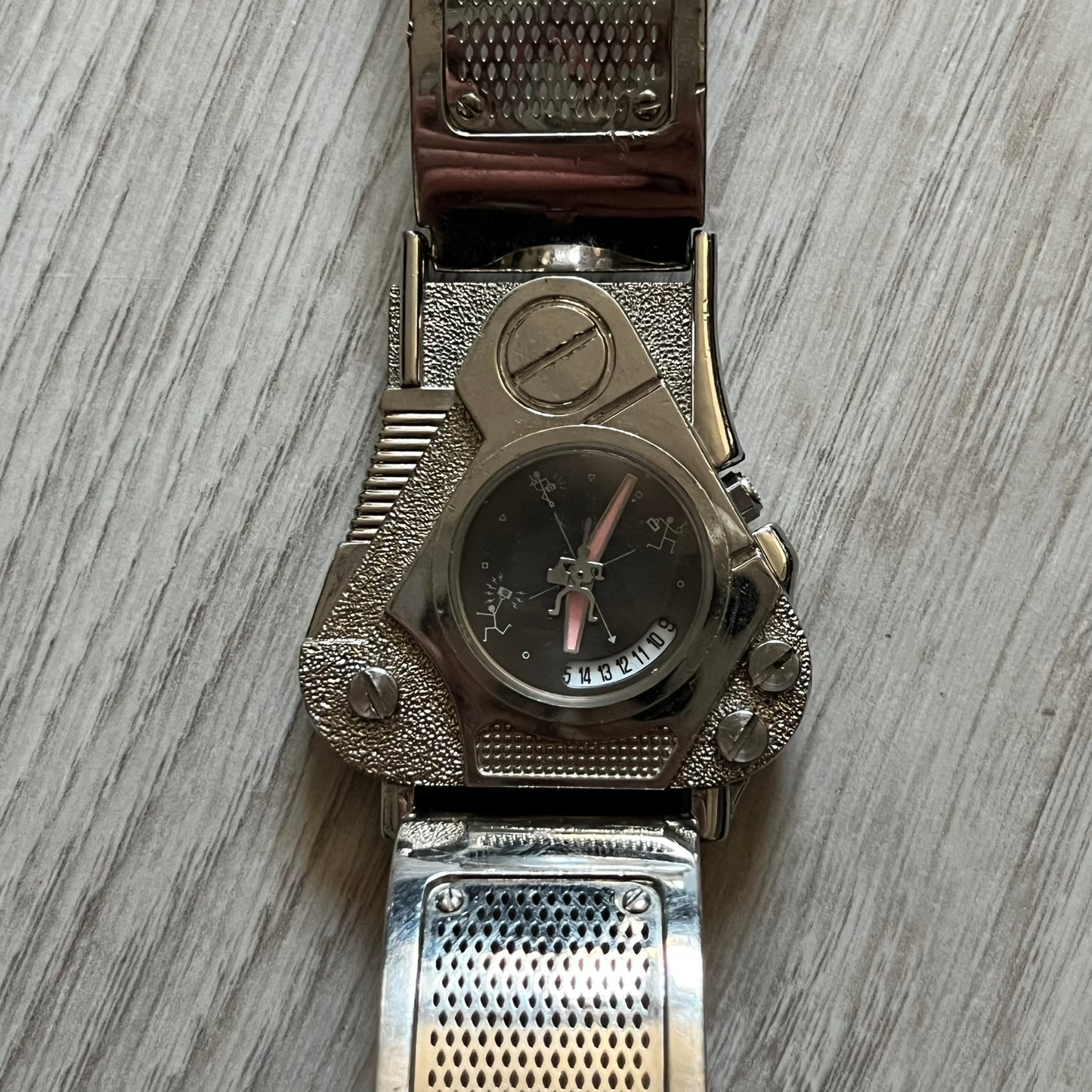 The Hi-Tek Alexander “The Engine” analog watch is a 1990s model made in England, designed with a sculptural case shaped like a motorcycle engine. It features a black dial with Alexander Hi-Tek figure logos in place of hour markers, pink hour and minute hands, and a Hi-Tek figure logo used as the seconds hand. A small sub-section at the bottom displays the day in numeric form, while visible screws and textured detailing are integrated into the engine-style face.&nbsp;The watch has an alloy case with a stainl