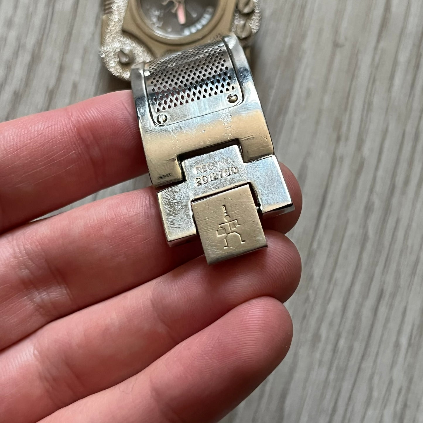 The Hi-Tek Alexander “The Engine” analog watch is a 1990s model made in England, designed with a sculptural case shaped like a motorcycle engine. It features a black dial with Alexander Hi-Tek figure logos in place of hour markers, pink hour and minute hands, and a Hi-Tek figure logo used as the seconds hand. A small sub-section at the bottom displays the day in numeric form, while visible screws and textured detailing are integrated into the engine-style face.&nbsp;The watch has an alloy case with a stainl