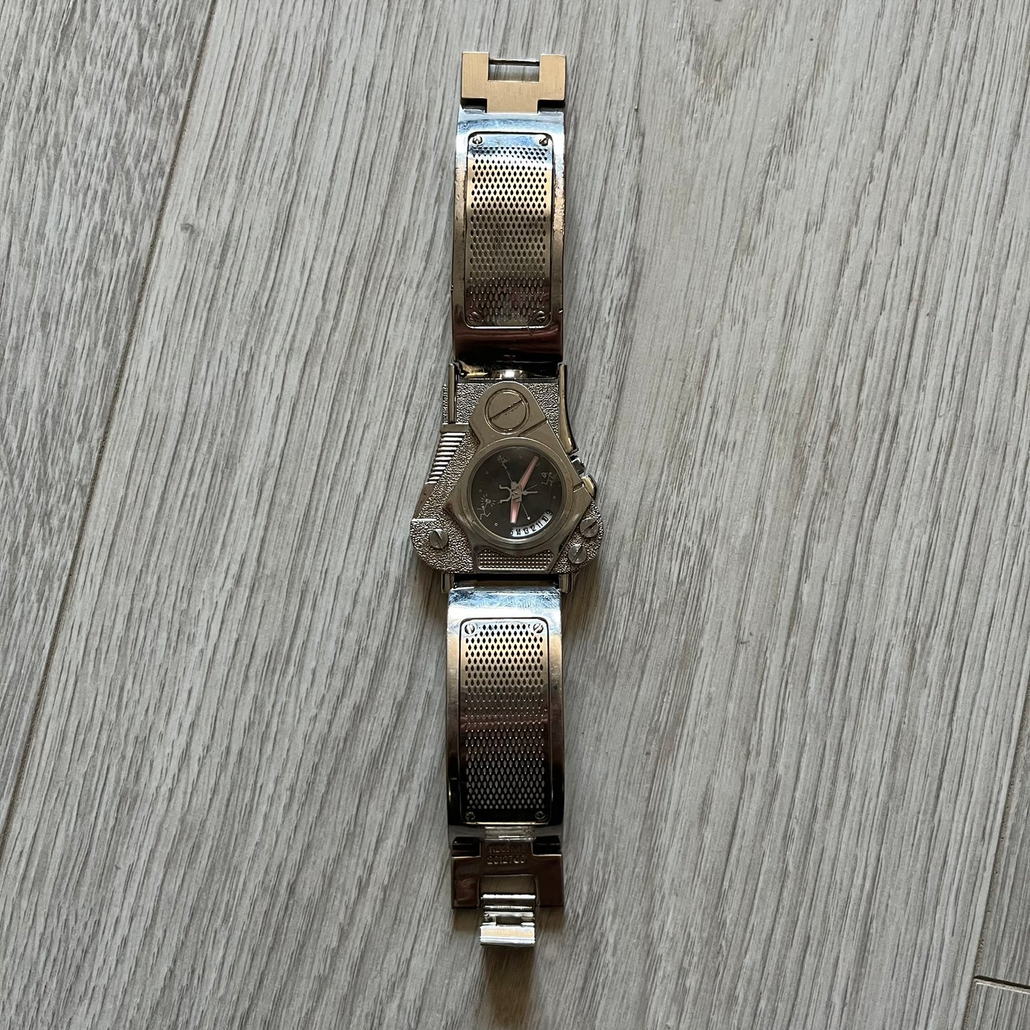 The Hi-Tek Alexander “The Engine” analog watch is a 1990s model made in England, designed with a sculptural case shaped like a motorcycle engine. It features a black dial with Alexander Hi-Tek figure logos in place of hour markers, pink hour and minute hands, and a Hi-Tek figure logo used as the seconds hand. A small sub-section at the bottom displays the day in numeric form, while visible screws and textured detailing are integrated into the engine-style face.&nbsp;The watch has an alloy case with a stainl