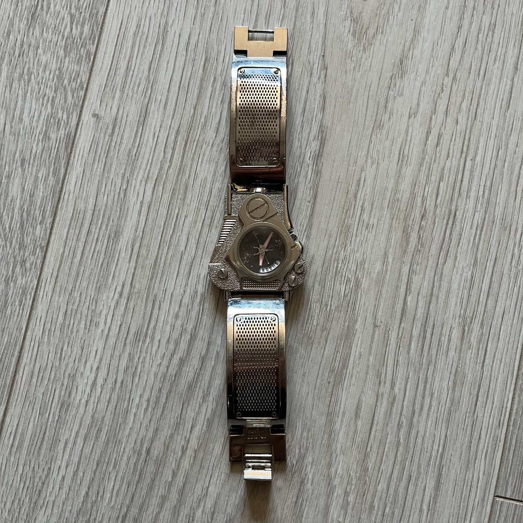 The Hi-Tek Alexander “The Engine” analog watch is a 1990s model made in England, designed with a sculptural case shaped like a motorcycle engine. It features a black dial with Alexander Hi-Tek figure logos in place of hour markers, pink hour and minute hands, and a Hi-Tek figure logo used as the seconds hand. A small sub-section at the bottom displays the day in numeric form, while visible screws and textured detailing are integrated into the engine-style face.&nbsp;The watch has an alloy case with a stainl
