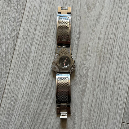 The Hi-Tek Alexander “The Engine” analog watch is a 1990s model made in England, designed with a sculptural case shaped like a motorcycle engine. It features a black dial with Alexander Hi-Tek figure logos in place of hour markers, pink hour and minute hands, and a Hi-Tek figure logo used as the seconds hand. A small sub-section at the bottom displays the day in numeric form, while visible screws and textured detailing are integrated into the engine-style face.&nbsp;The watch has an alloy case with a stainl