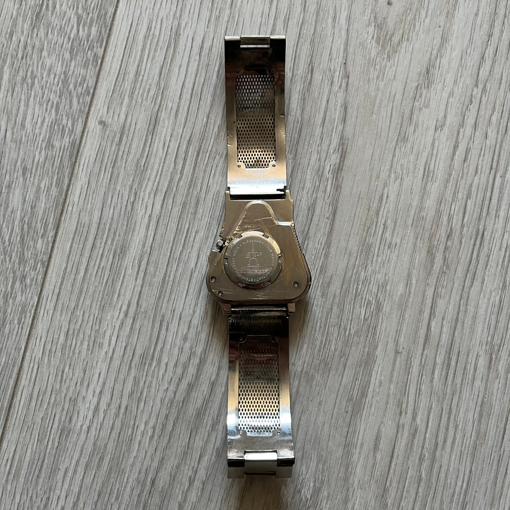 The Hi-Tek Alexander “The Engine” analog watch is a 1990s model made in England, designed with a sculptural case shaped like a motorcycle engine. It features a black dial with Alexander Hi-Tek figure logos in place of hour markers, pink hour and minute hands, and a Hi-Tek figure logo used as the seconds hand. A small sub-section at the bottom displays the day in numeric form, while visible screws and textured detailing are integrated into the engine-style face.&nbsp;The watch has an alloy case with a stainl