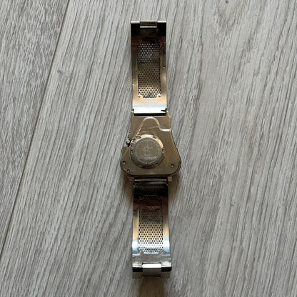 The Hi-Tek Alexander “The Engine” analog watch is a 1990s model made in England, designed with a sculptural case shaped like a motorcycle engine. It features a black dial with Alexander Hi-Tek figure logos in place of hour markers, pink hour and minute hands, and a Hi-Tek figure logo used as the seconds hand. A small sub-section at the bottom displays the day in numeric form, while visible screws and textured detailing are integrated into the engine-style face.&nbsp;The watch has an alloy case with a stainl