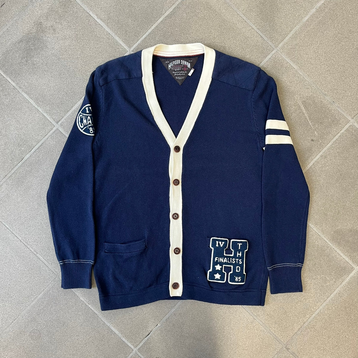 Hilfiger Denim 2012 Navy Varsity Cardigan is a navy cardigan with a white collar/hem trim and brown buttons, featuring two front pockets. It includes a large fuzzy "H" patch with text, two white straps on one sleeve, and a fuzzy white and navy "IV CHAMPS '85" patch.