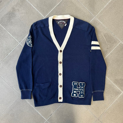 Hilfiger Denim 2012 Navy Varsity Cardigan is a navy cardigan with a white collar/hem trim and brown buttons, featuring two front pockets. It includes a large fuzzy "H" patch with text, two white straps on one sleeve, and a fuzzy white and navy "IV CHAMPS '85" patch.