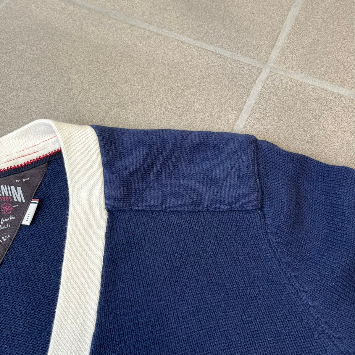 Hilfiger Denim 2012 Navy Varsity Cardigan is a navy cardigan with a white collar/hem trim and brown buttons, featuring two front pockets. It includes a large fuzzy "H" patch with text, two white straps on one sleeve, and a fuzzy white and navy "IV CHAMPS '85" patch.