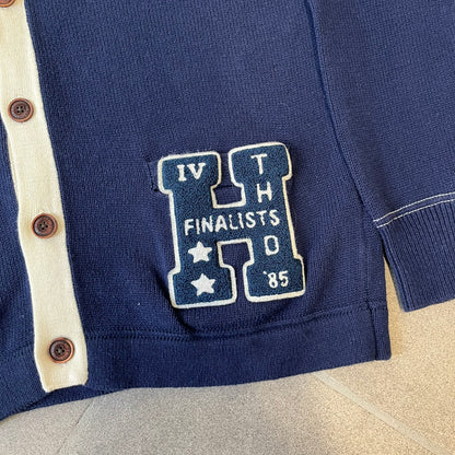 Hilfiger Denim 2012 Navy Varsity Cardigan is a navy cardigan with a white collar/hem trim and brown buttons, featuring two front pockets. It includes a large fuzzy "H" patch with text, two white straps on one sleeve, and a fuzzy white and navy "IV CHAMPS '85" patch.