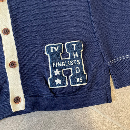 Hilfiger Denim 2012 Navy Varsity Cardigan is a navy cardigan with a white collar/hem trim and brown buttons, featuring two front pockets. It includes a large fuzzy "H" patch with text, two white straps on one sleeve, and a fuzzy white and navy "IV CHAMPS '85" patch.