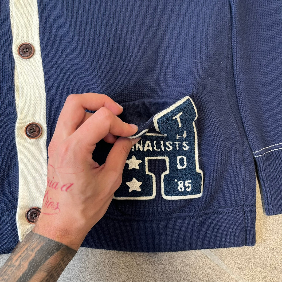 Hilfiger Denim 2012 Navy Varsity Cardigan is a navy cardigan with a white collar/hem trim and brown buttons, featuring two front pockets. It includes a large fuzzy "H" patch with text, two white straps on one sleeve, and a fuzzy white and navy "IV CHAMPS '85" patch.