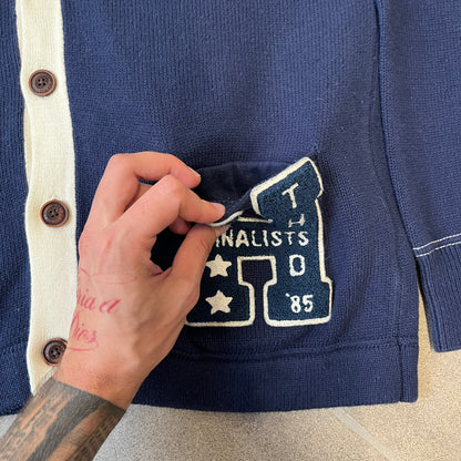 Hilfiger Denim 2012 Navy Varsity Cardigan is a navy cardigan with a white collar/hem trim and brown buttons, featuring two front pockets. It includes a large fuzzy "H" patch with text, two white straps on one sleeve, and a fuzzy white and navy "IV CHAMPS '85" patch.