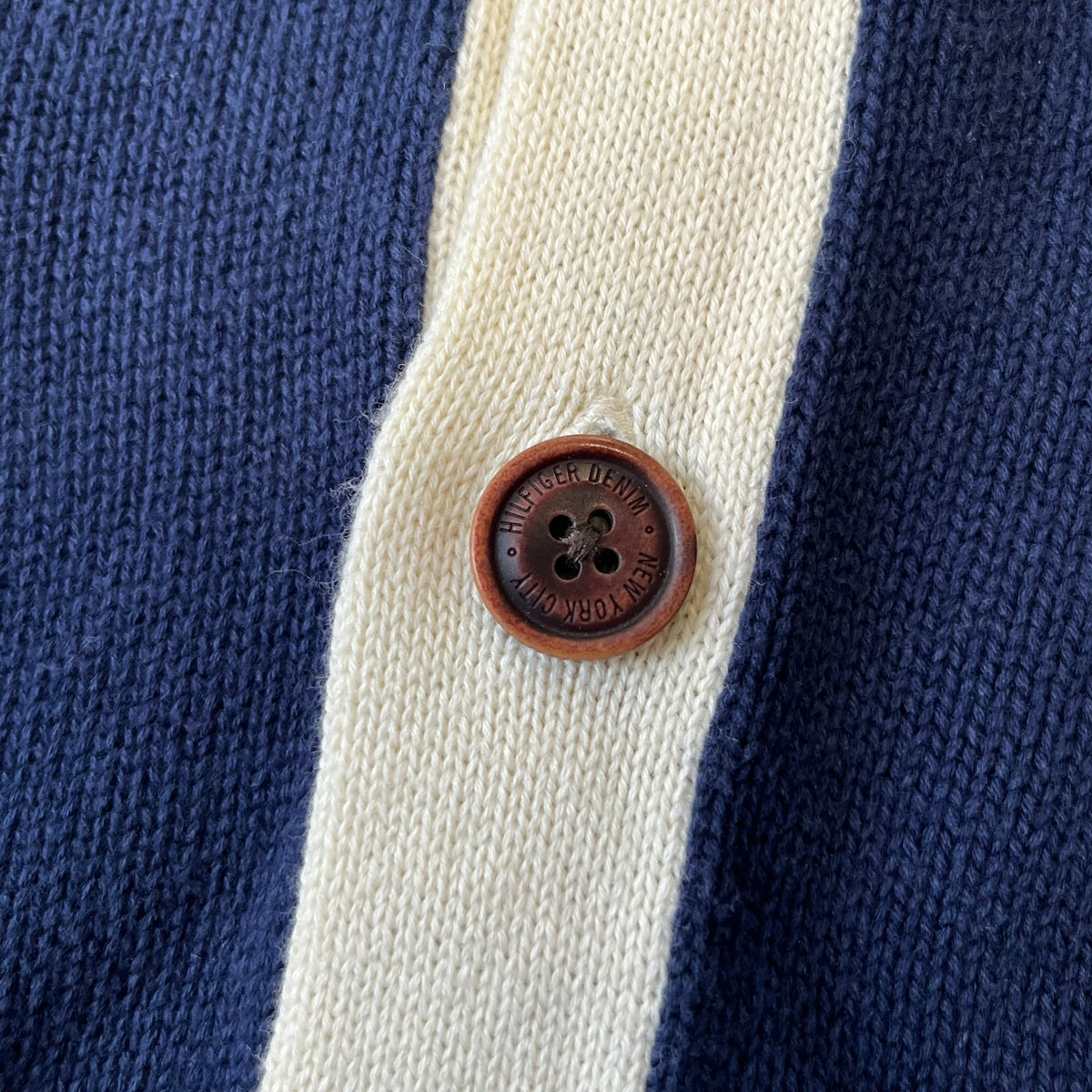 Hilfiger Denim 2012 Navy Varsity Cardigan is a navy cardigan with a white collar/hem trim and brown buttons, featuring two front pockets. It includes a large fuzzy "H" patch with text, two white straps on one sleeve, and a fuzzy white and navy "IV CHAMPS '85" patch.