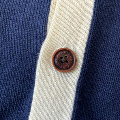 Hilfiger Denim 2012 Navy Varsity Cardigan is a navy cardigan with a white collar/hem trim and brown buttons, featuring two front pockets. It includes a large fuzzy "H" patch with text, two white straps on one sleeve, and a fuzzy white and navy "IV CHAMPS '85" patch.
