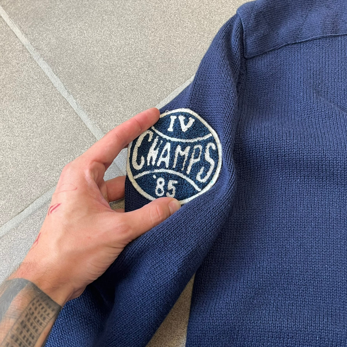 Hilfiger Denim 2012 Navy Varsity Cardigan is a navy cardigan with a white collar/hem trim and brown buttons, featuring two front pockets. It includes a large fuzzy "H" patch with text, two white straps on one sleeve, and a fuzzy white and navy "IV CHAMPS '85" patch.