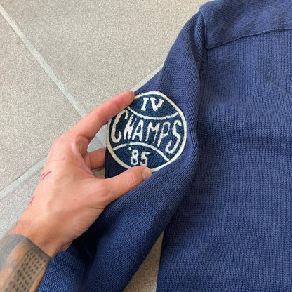 Hilfiger Denim 2012 Navy Varsity Cardigan is a navy cardigan with a white collar/hem trim and brown buttons, featuring two front pockets. It includes a large fuzzy "H" patch with text, two white straps on one sleeve, and a fuzzy white and navy "IV CHAMPS '85" patch.
