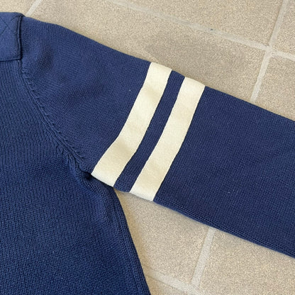 Hilfiger Denim 2012 Navy Varsity Cardigan is a navy cardigan with a white collar/hem trim and brown buttons, featuring two front pockets. It includes a large fuzzy "H" patch with text, two white straps on one sleeve, and a fuzzy white and navy "IV CHAMPS '85" patch.