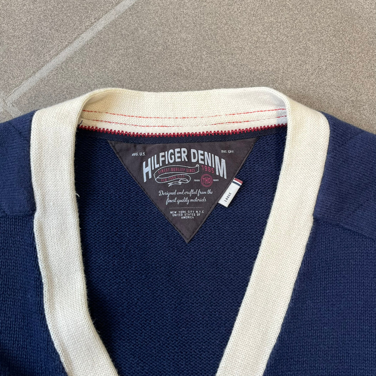 Hilfiger Denim 2012 Navy Varsity Cardigan is a navy cardigan with a white collar/hem trim and brown buttons, featuring two front pockets. It includes a large fuzzy "H" patch with text, two white straps on one sleeve, and a fuzzy white and navy "IV CHAMPS '85" patch.