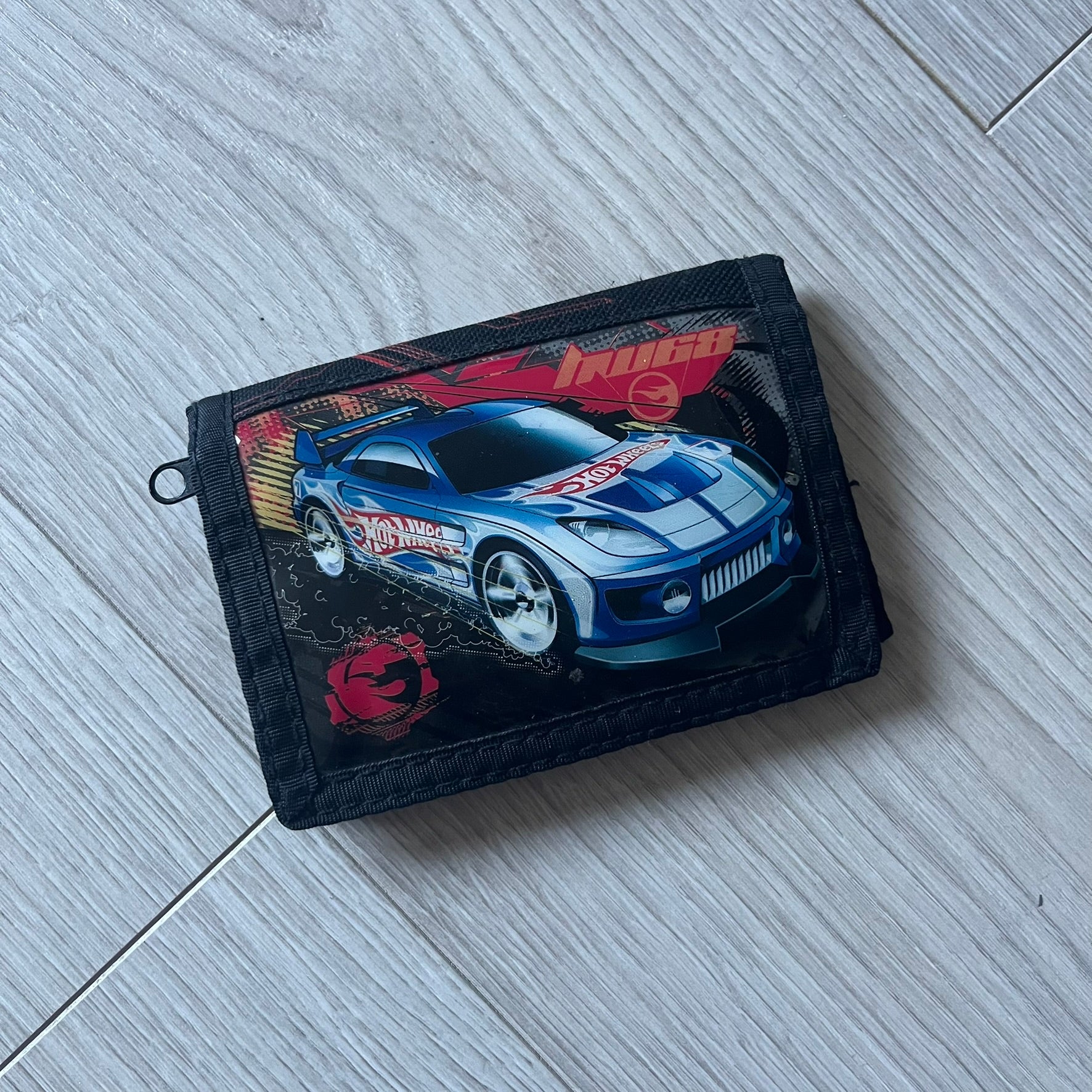 2008 Hot Wheels 24/Seven wallet in black with a detailed blue 24/Seven Car graphic under a protective plastic laminate. Includes Hot Wheels logo detailing, Mattel tags, and plenty of compartments for storage. A functional piece with nostalgic collector appeal.