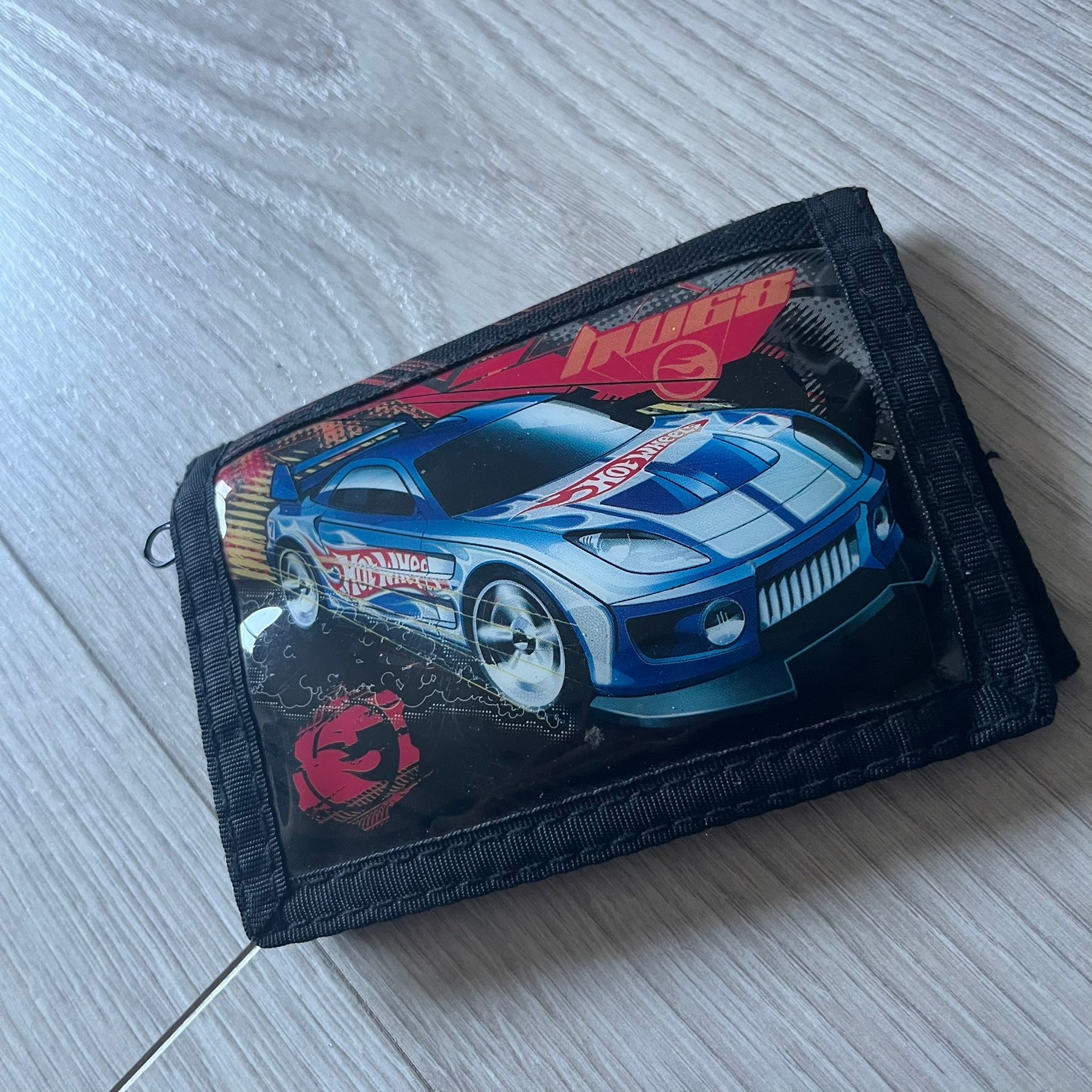 2008 Hot Wheels 24/Seven wallet in black with a detailed blue 24/Seven Car graphic under a protective plastic laminate. Includes Hot Wheels logo detailing, Mattel tags, and plenty of compartments for storage. A functional piece with nostalgic collector appeal.