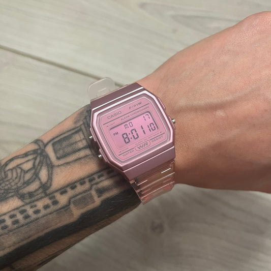 A digital Casio watch in pink color, displayed on a person's wrist against a wooden background.