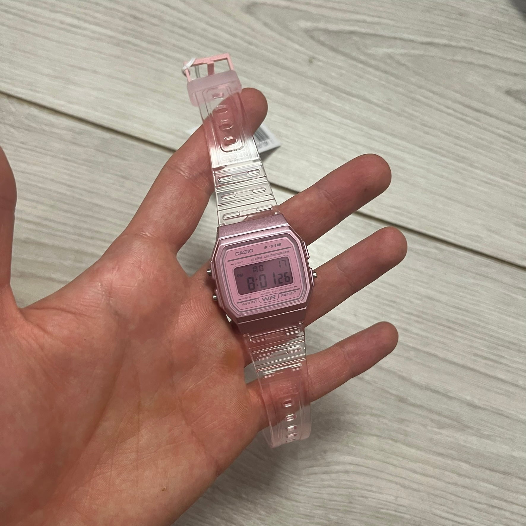 Hand holding a pink digital watch with clear strap against a wooden background.