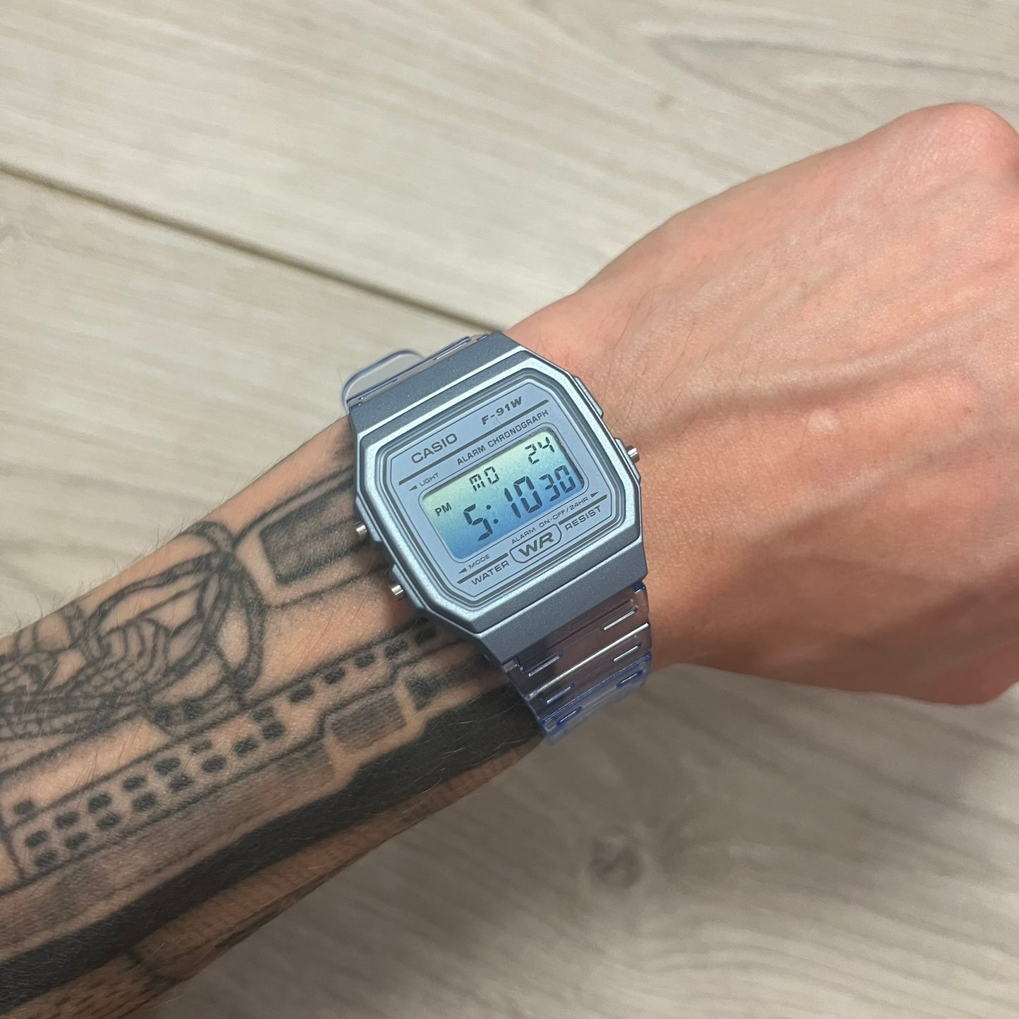 A digital Casio watch in blue color, displayed on a person's wrist against a wooden background.