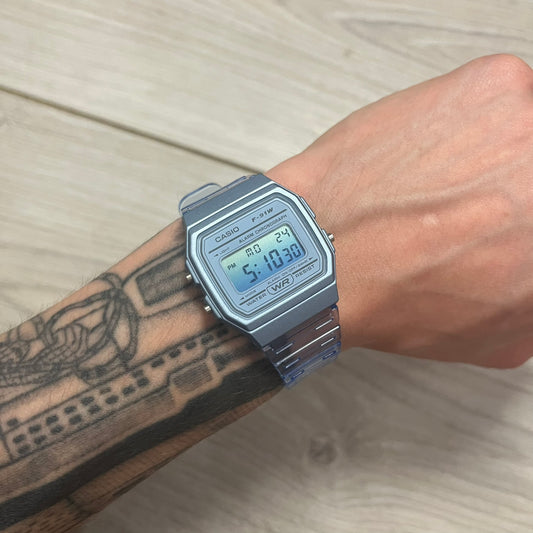 A digital Casio watch in blue color, displayed on a person's wrist against a wooden background.