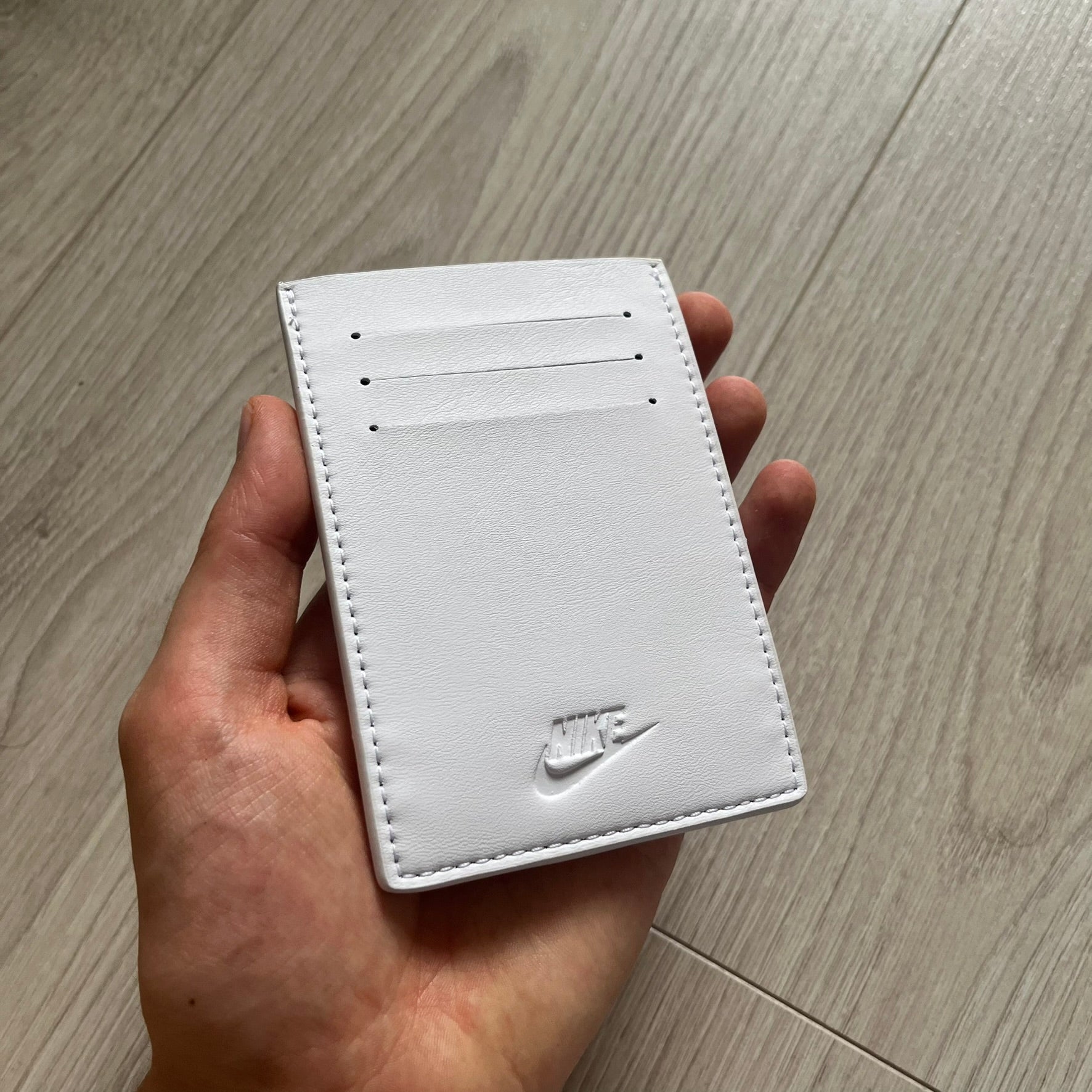 White leather cardholder with Nike logo held in a hand on a wooden floor.