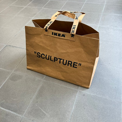Ikea x Virgil Abloh Markerad "Sculpture" Carrier Bag from FW19 reimagines the iconic blue Frakta tote in reinforced brown paper. Embellished with Abloh’s signature quotation marks, the “SCULPTURE” print transforms a utilitarian object into conceptual design. A collectible symbol of functional art and everyday irony.