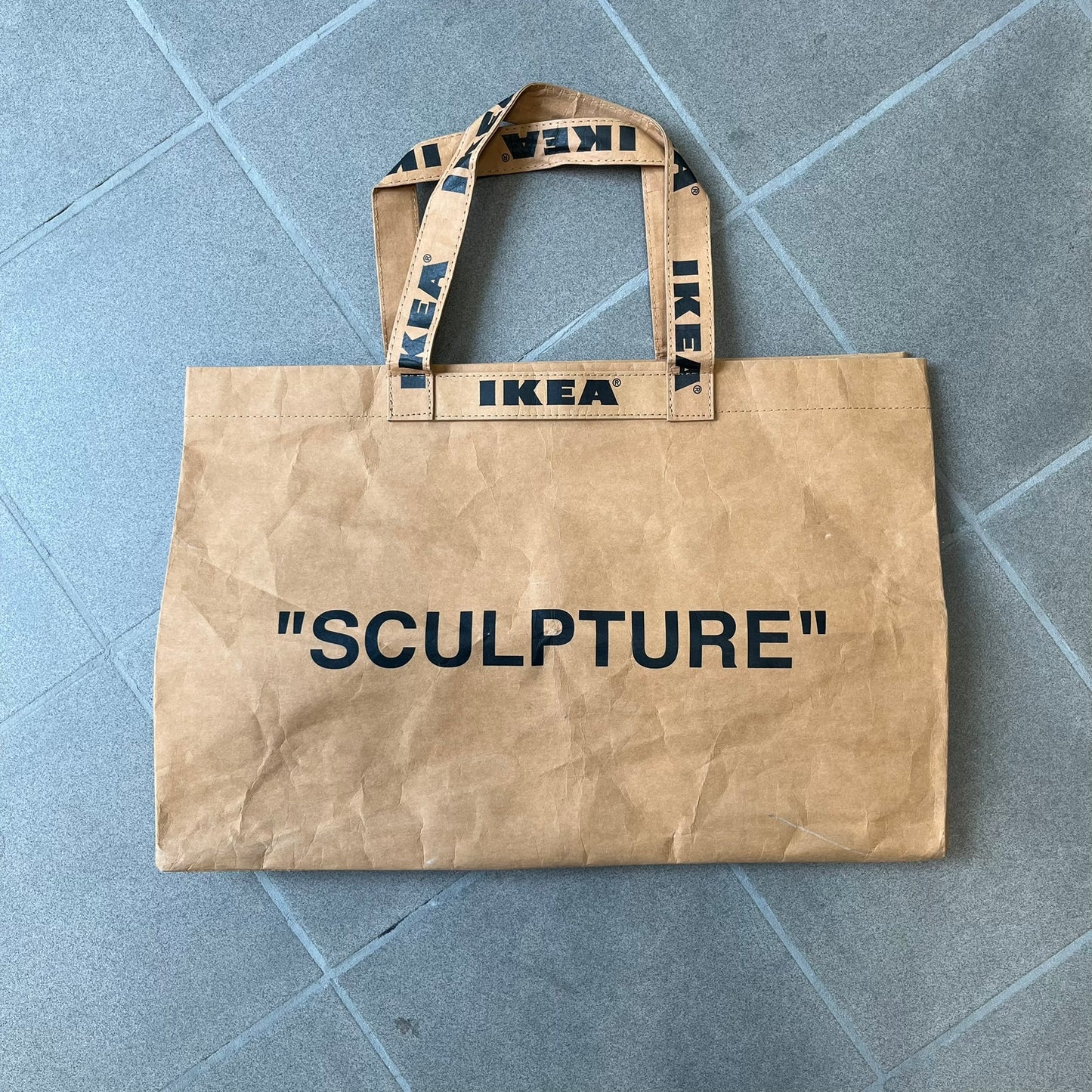 Ikea x Virgil Abloh Markerad "Sculpture" Carrier Bag from FW19 reimagines the iconic blue Frakta tote in reinforced brown paper. Embellished with Abloh’s signature quotation marks, the “SCULPTURE” print transforms a utilitarian object into conceptual design. A collectible symbol of functional art and everyday irony.