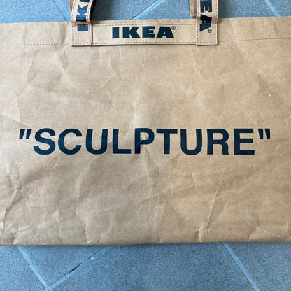 Ikea x Virgil Abloh Markerad "Sculpture" Carrier Bag from FW19 reimagines the iconic blue Frakta tote in reinforced brown paper. Embellished with Abloh’s signature quotation marks, the “SCULPTURE” print transforms a utilitarian object into conceptual design. A collectible symbol of functional art and everyday irony.