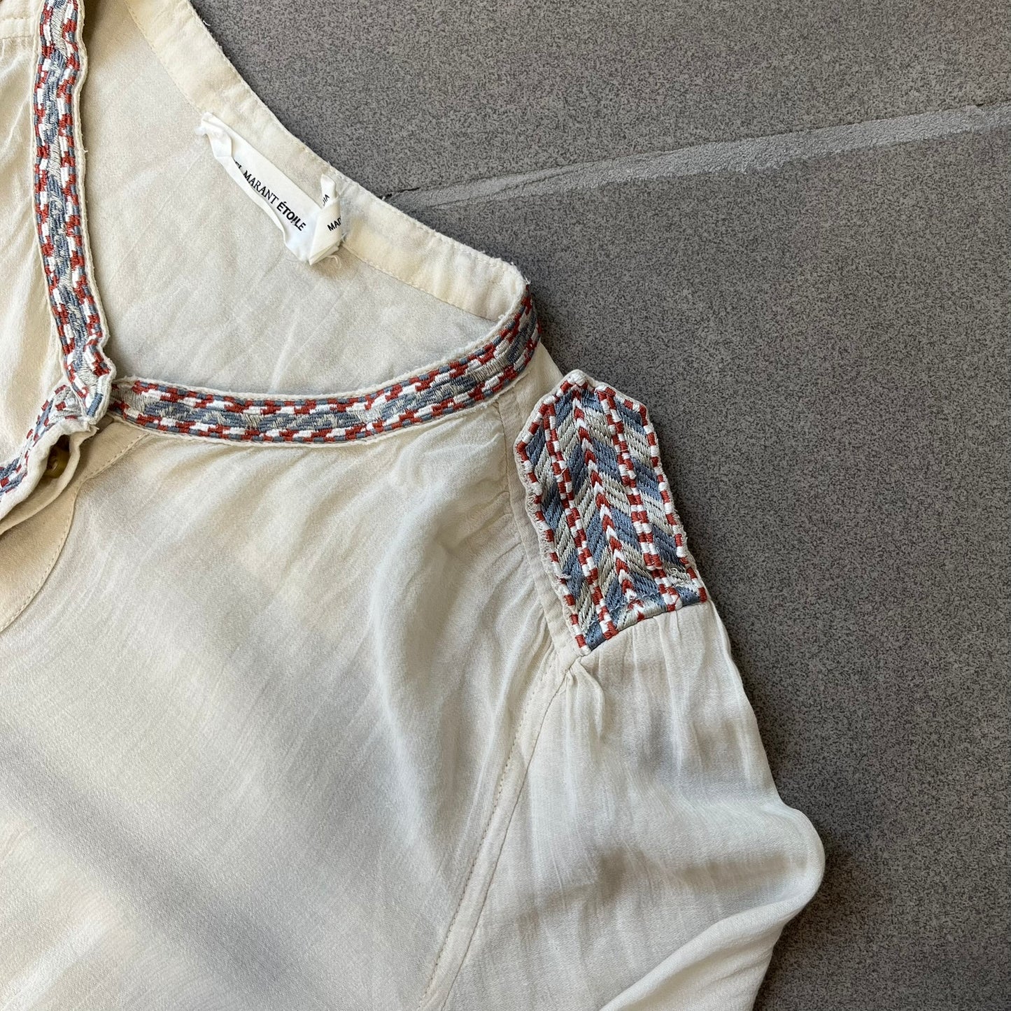 Isabel Marant Étoile Cream Embroidered Blouse features a lightweight, semi-sheer cream fabric with multicolored embroidery along the front button placket, collar, and shoulder details. It has a button-up front and one chest pocket.