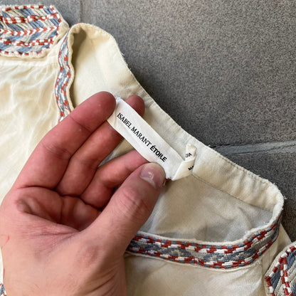 Isabel Marant Étoile Cream Embroidered Blouse features a lightweight, semi-sheer cream fabric with multicolored embroidery along the front button placket, collar, and shoulder details. It has a button-up front and one chest pocket.