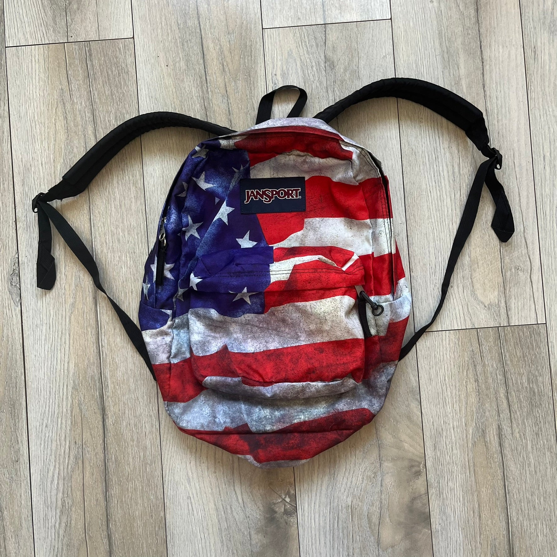 JanSport High Stakes Multi Grunge USA Backpack (JS00TRS70GM) is a backpack featuring an all-over distressed American flag design across the front and side panels. The flag graphic has a deliberately worn and faded appearance, giving it a vintage grunge look. The back panel and shoulder straps are finished in black, while the front includes the classic JanSport logo patch.