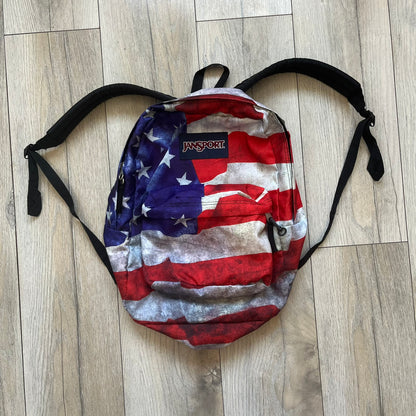 JanSport High Stakes Multi Grunge USA Backpack (JS00TRS70GM) is a backpack featuring an all-over distressed American flag design across the front and side panels. The flag graphic has a deliberately worn and faded appearance, giving it a vintage grunge look. The back panel and shoulder straps are finished in black, while the front includes the classic JanSport logo patch.