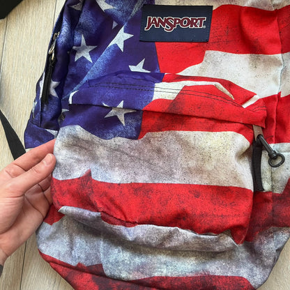 JanSport High Stakes Multi Grunge USA Backpack (JS00TRS70GM) is a backpack featuring an all-over distressed American flag design across the front and side panels. The flag graphic has a deliberately worn and faded appearance, giving it a vintage grunge look. The back panel and shoulder straps are finished in black, while the front includes the classic JanSport logo patch.