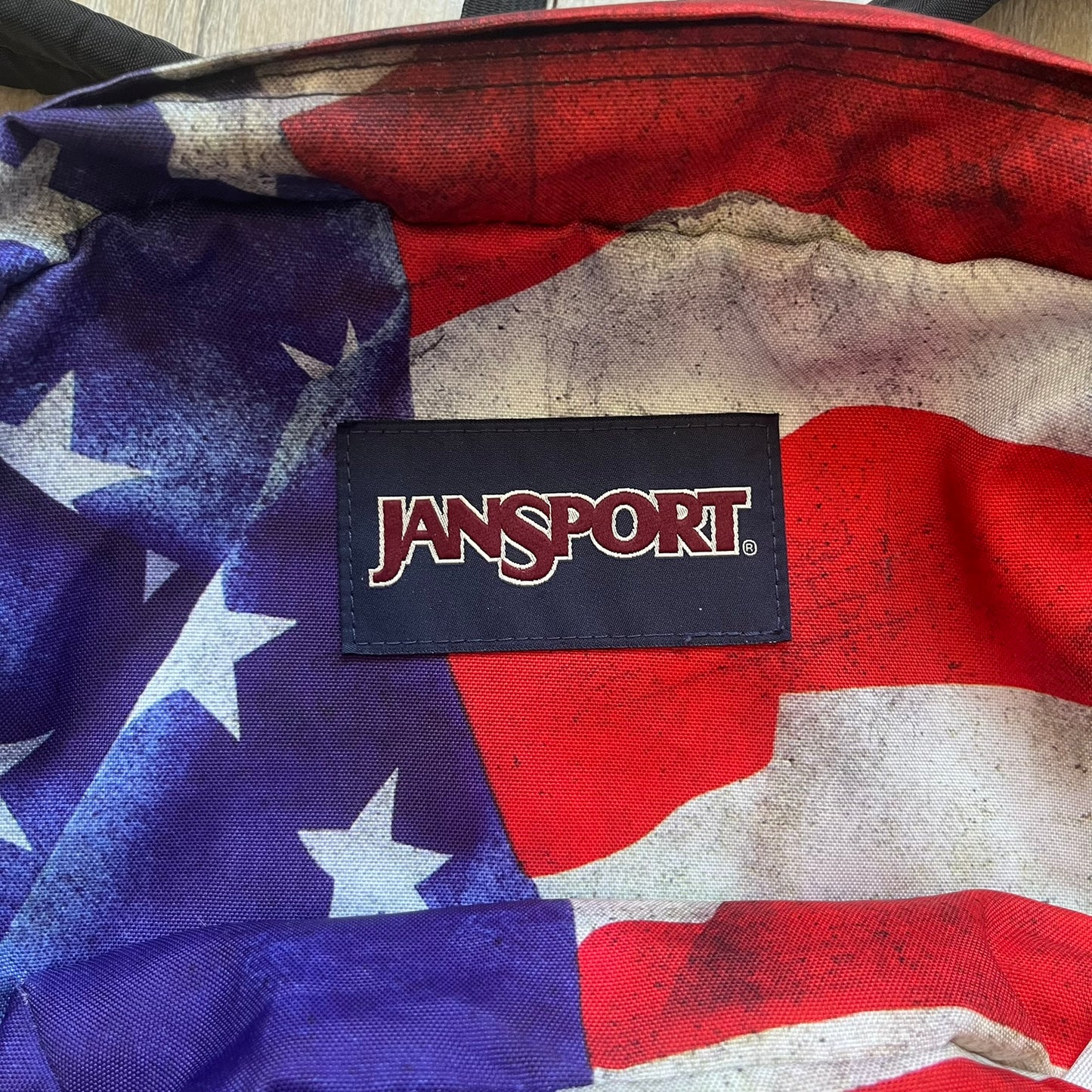 JanSport High Stakes Multi Grunge USA Backpack (JS00TRS70GM) is a backpack featuring an all-over distressed American flag design across the front and side panels. The flag graphic has a deliberately worn and faded appearance, giving it a vintage grunge look. The back panel and shoulder straps are finished in black, while the front includes the classic JanSport logo patch.