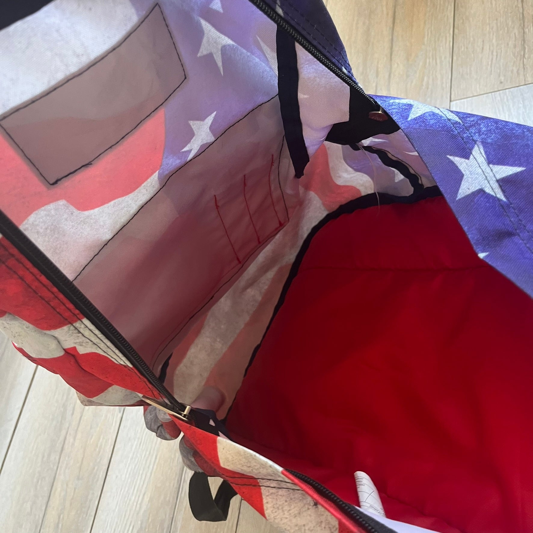 JanSport High Stakes Multi Grunge USA Backpack (JS00TRS70GM) is a backpack featuring an all-over distressed American flag design across the front and side panels. The flag graphic has a deliberately worn and faded appearance, giving it a vintage grunge look. The back panel and shoulder straps are finished in black, while the front includes the classic JanSport logo patch.
