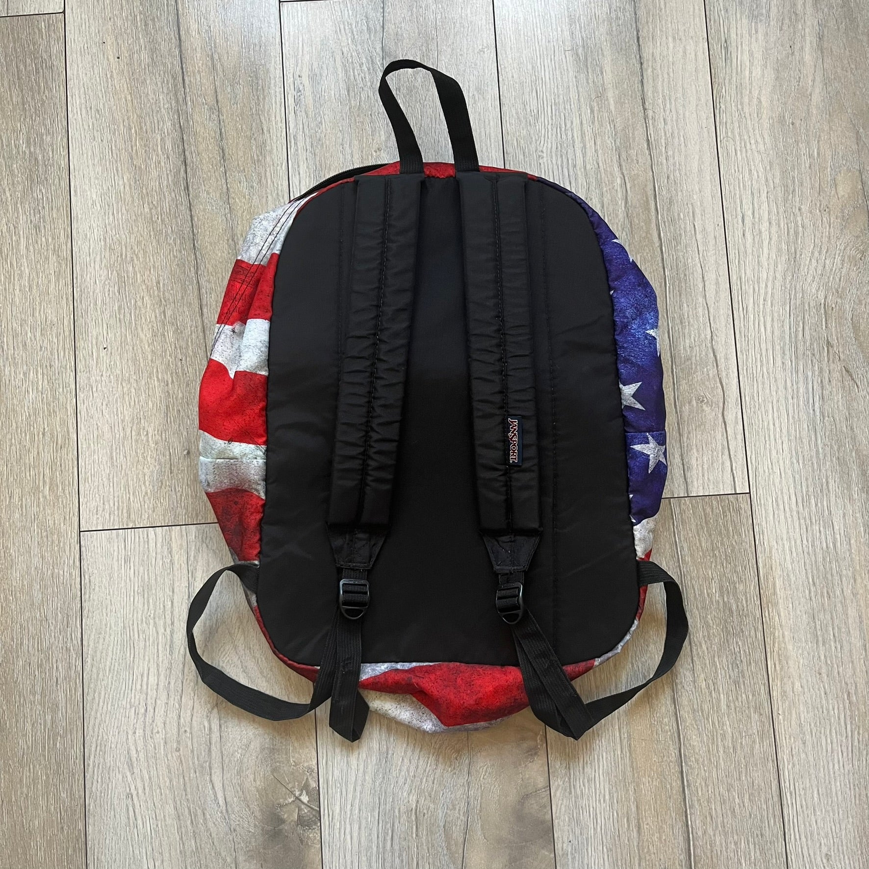JanSport High Stakes Multi Grunge USA Backpack (JS00TRS70GM) is a backpack featuring an all-over distressed American flag design across the front and side panels. The flag graphic has a deliberately worn and faded appearance, giving it a vintage grunge look. The back panel and shoulder straps are finished in black, while the front includes the classic JanSport logo patch.