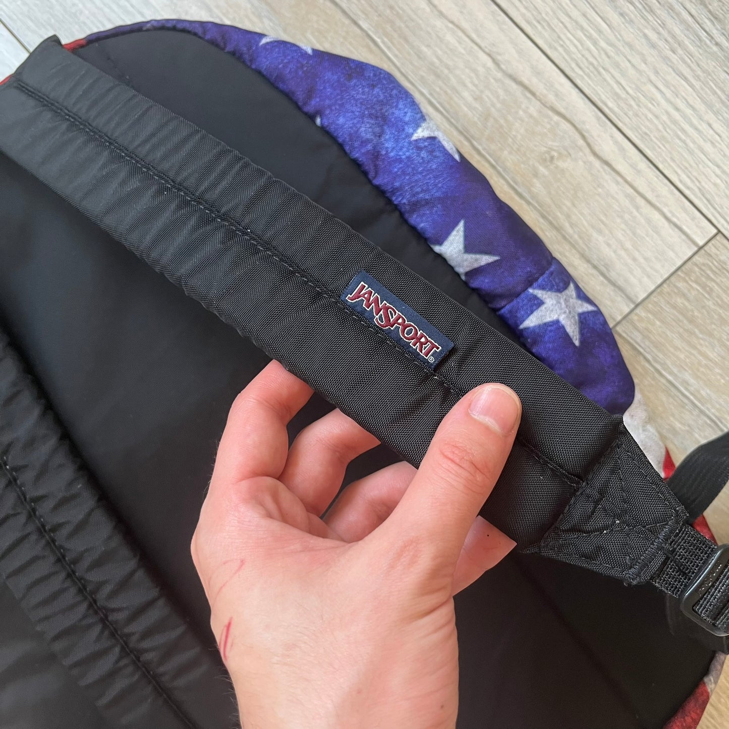 JanSport High Stakes Multi Grunge USA Backpack (JS00TRS70GM) is a backpack featuring an all-over distressed American flag design across the front and side panels. The flag graphic has a deliberately worn and faded appearance, giving it a vintage grunge look. The back panel and shoulder straps are finished in black, while the front includes the classic JanSport logo patch.