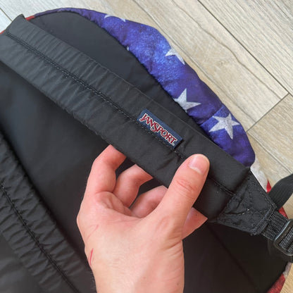 JanSport High Stakes Multi Grunge USA Backpack (JS00TRS70GM) is a backpack featuring an all-over distressed American flag design across the front and side panels. The flag graphic has a deliberately worn and faded appearance, giving it a vintage grunge look. The back panel and shoulder straps are finished in black, while the front includes the classic JanSport logo patch.