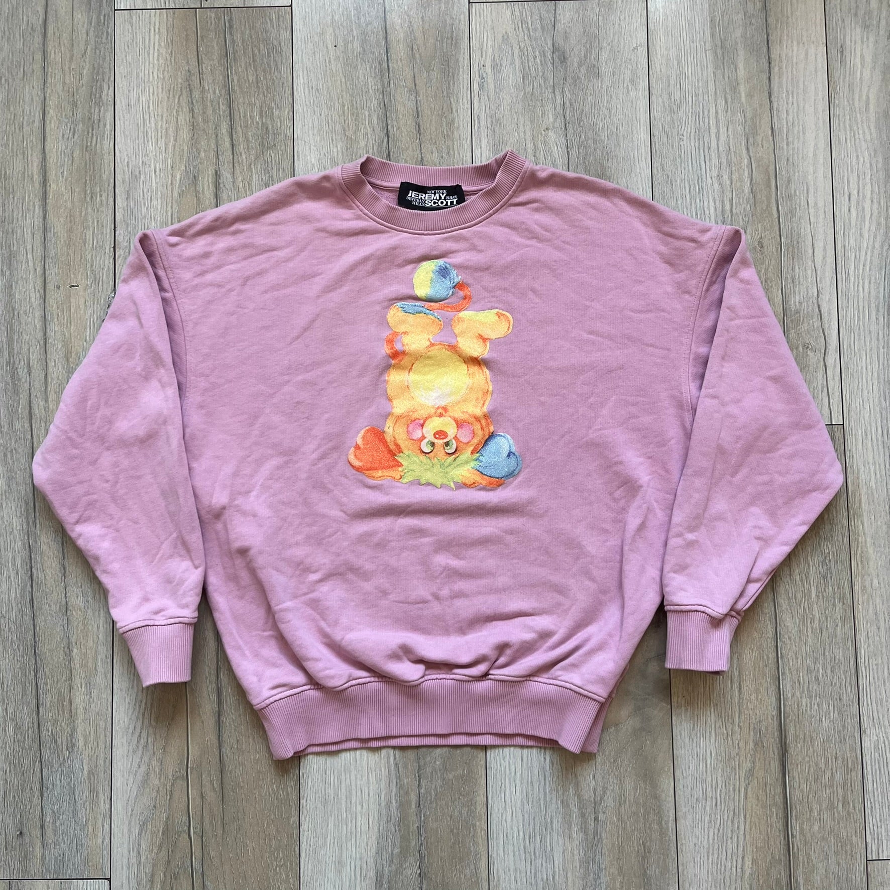 Jeremy Scott 2018 Popples Pink Crewneck is a pink sweatshirt featuring a front graphic of an upside-down orange Popples character with green hair and blue/red heart-shaped ears, finished with a glittery, shiny application. The upper back includes a rubberized orange “Jeremy Scott Paris” logo.

