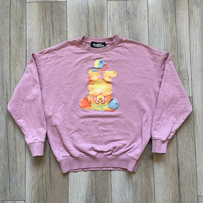 Jeremy Scott 2018 Popples Pink Crewneck is a pink sweatshirt featuring a front graphic of an upside-down orange Popples character with green hair and blue/red heart-shaped ears, finished with a glittery, shiny application. The upper back includes a rubberized orange “Jeremy Scott Paris” logo.
