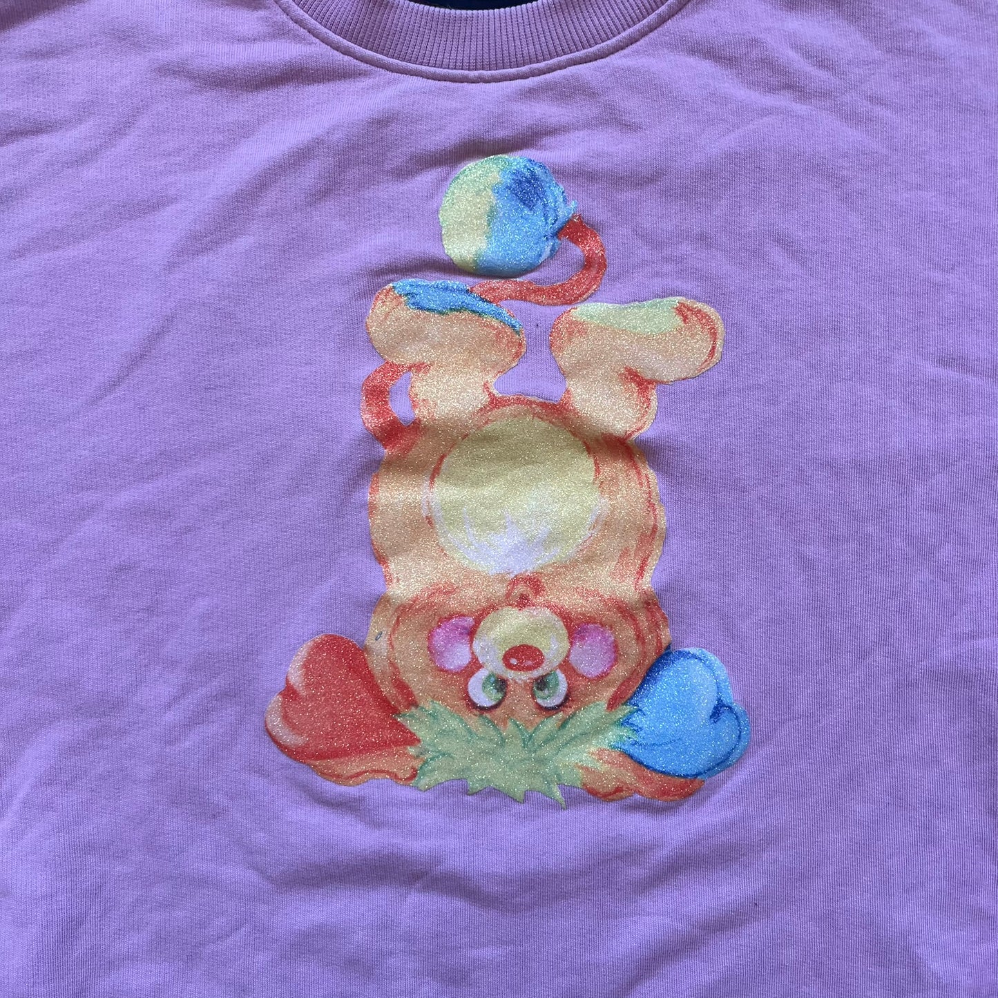 Jeremy Scott 2018 Popples Pink Crewneck is a pink sweatshirt featuring a front graphic of an upside-down orange Popples character with green hair and blue/red heart-shaped ears, finished with a glittery, shiny application. The upper back includes a rubberized orange “Jeremy Scott Paris” logo.
