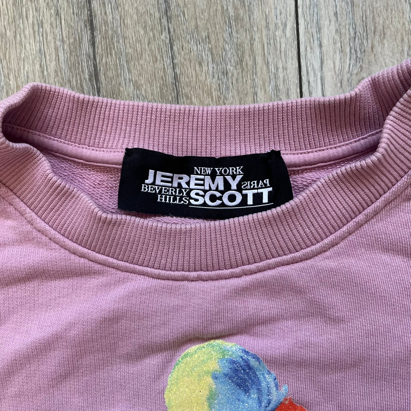 Jeremy Scott 2018 Popples Pink Crewneck is a pink sweatshirt featuring a front graphic of an upside-down orange Popples character with green hair and blue/red heart-shaped ears, finished with a glittery, shiny application. The upper back includes a rubberized orange “Jeremy Scott Paris” logo.
