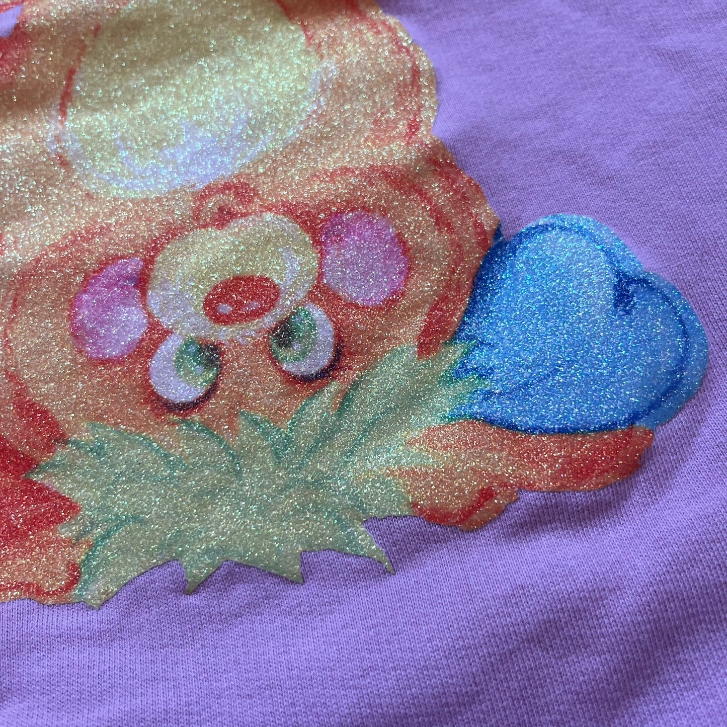 Jeremy Scott 2018 Popples Pink Crewneck is a pink sweatshirt featuring a front graphic of an upside-down orange Popples character with green hair and blue/red heart-shaped ears, finished with a glittery, shiny application. The upper back includes a rubberized orange “Jeremy Scott Paris” logo.
